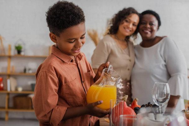 Smiling african american boy pouring orange juice near blurred mother and granny during thanksgiving dinner  - 写真・画像
