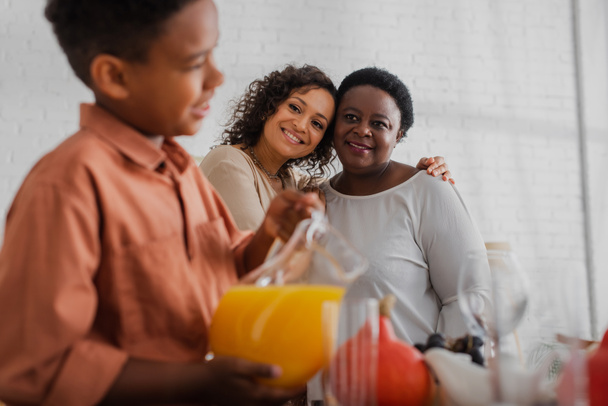 glimlachende Afro-Amerikaanse vrouwen kijken naar wazig kind met sinaasappelsap in de buurt van Thanksgiving diner  - Foto, afbeelding