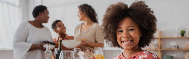 Smiling african american girl looking at camera near blurred family and thanksgiving dinner, banner  - Foto, immagini
