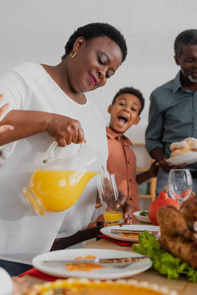 Африканская американская бабушка наливает апельсиновый сок рядом с размытой семьей и праздничным ужином  - Фото, изображение