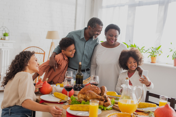 African american girl holding smartphone near family and thanksgiving dinner  - Photo, Image