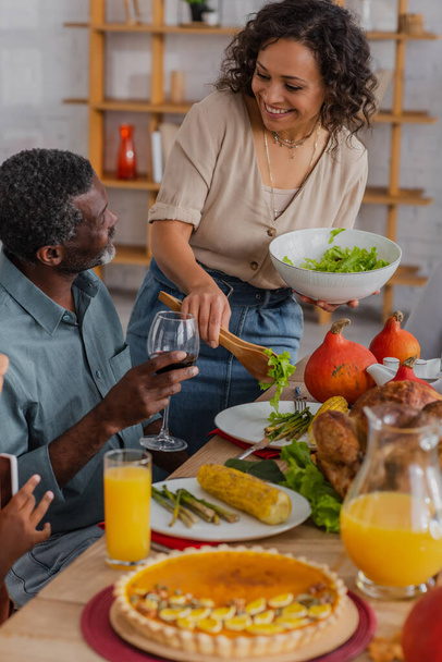 Smiling african american woman pouring salad near father with wine and thanksgiving celebration  - Photo, Image