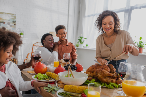 Smiling african american woman cutting turkey near family and thanksgiving dinner - Фото, зображення