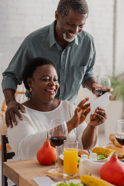 African american woman using smartphone near husband with wine and thanksgiving dinner  - 写真・画像