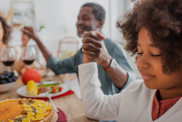 selective focus of african american girl with closed eyes praying with blurred grandpa and mom on thanksgiving dinner - Photo, Image