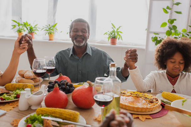 middle aged african american man with closed eyes praying with family during thanksgiving dinner - Фото, зображення