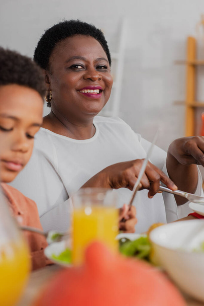 joyful african american woman looking at camera while having thanksgiving dinner near blurred grandson - Photo, image