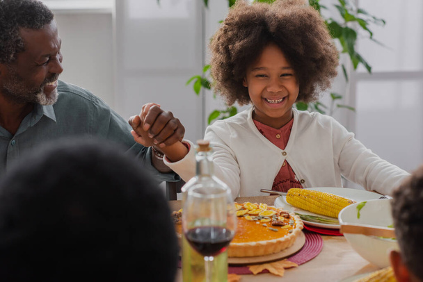 happy african american girl holding hands with grandfather during pray before thanksgiving dinner with blurred family - Photo, Image