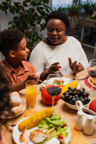 mature african american woman talking to grandson and blurred daughter near table served with thanksgiving dinner - Fotó, kép