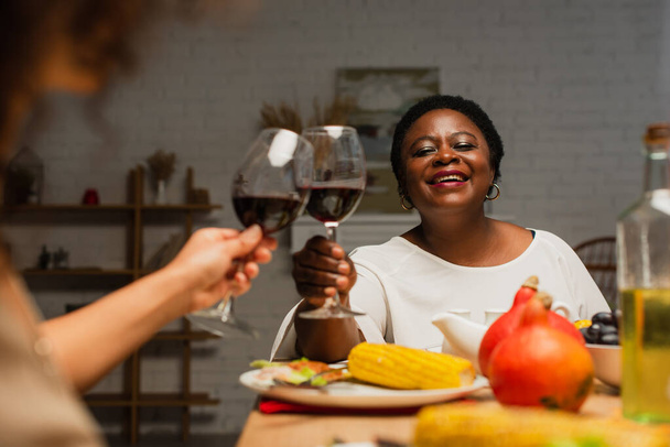 joyful african american woman clinking glasses of red wine with blurred daughter on thanksgiving dinner - Foto, Imagem