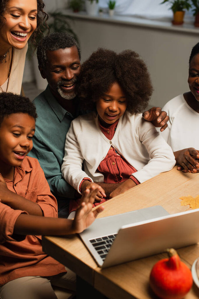 Africain américain garçon agitant la main près d'un ordinateur portable et la famille souriante lors d'un appel vidéo le jour de l'Action de grâces - Photo, image