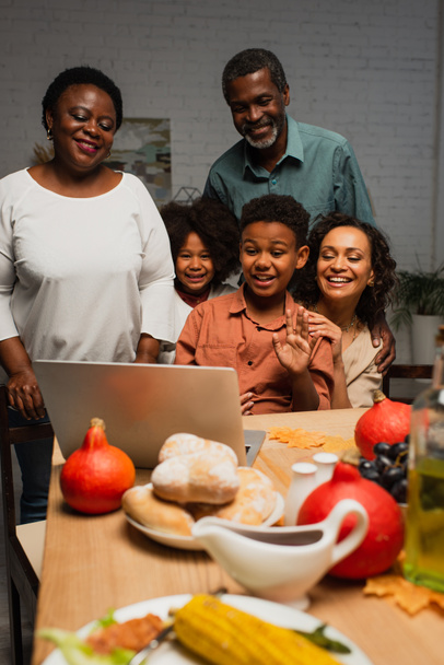 cheerful african american boy waving hand during video call near family and served dinner - Photo, Image
