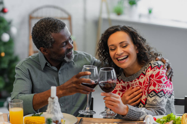 laughing african american woman clinking glasses of red wine with dad during christmas dinner - Fotó, kép