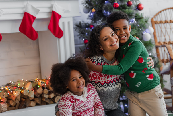 happy african american woman with kids hugging near blurred christmas tree and decorated fireplace - Foto, immagini