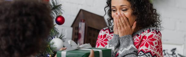 femme afro-américaine étonnée couvrant la bouche avec les mains près de fille floue avec cadeau de Noël, bannière - Photo, image