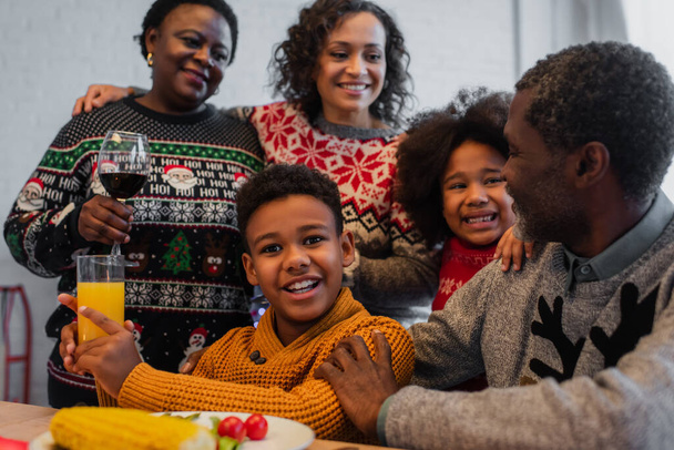 niño afroamericano feliz con jugo de naranja cerca de la familia en la cena de Navidad - Foto, imagen