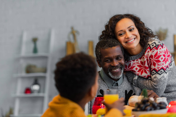 cheerful african american father and daughter looking at blurred boy during thanksgiving dinner - 写真・画像
