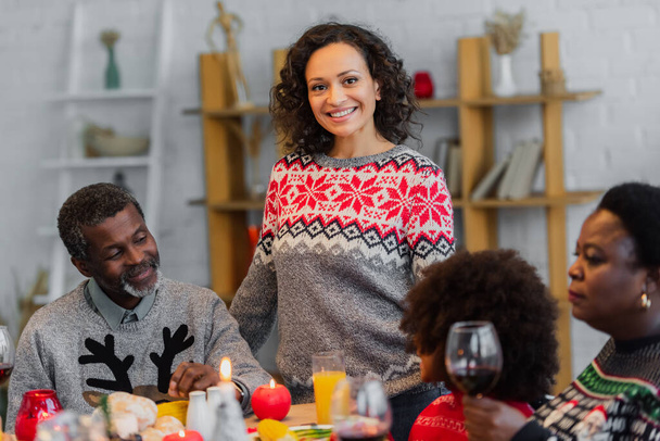 heureuse femme afro-américaine souriant à la caméra pendant le dîner de Noël avec les parents et la fille - Photo, image