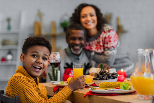 happy african american boy looking at camera during thanksgiving dinner with blurred family - Fotó, kép