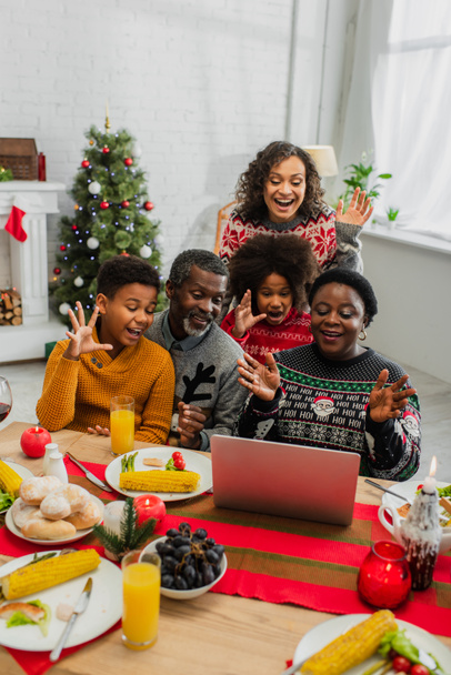 ενθουσιασμένοι αφρικάνικη αμερικανική οικογένεια χαιρετώντας τα χέρια, ενώ έχουν βιντεοκλήση κατά τη διάρκεια των Χριστουγέννων δείπνο - Φωτογραφία, εικόνα