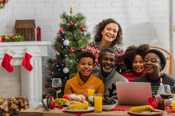 familia afroamericana sonriendo cerca del ordenador portátil en la mesa con la cena de Navidad - Foto, Imagen
