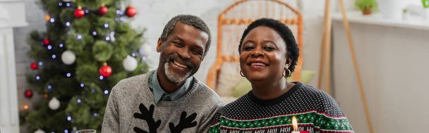 joyeux couple afro-américain d'âge moyen souriant à la caméra près de l'arbre de Noël flou, bannière - Photo, image