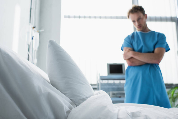 lit d'hôpital avec literie blanche près homme triste en robe de patient debout avec les bras croisés sur fond flou  - Photo, image