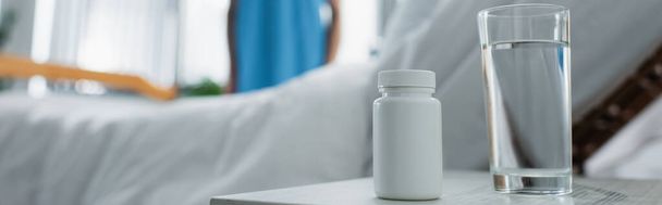 láhev s léky a sklenicí vody na nočním stolku u rozmazaného pacienta v nemocnici, banner - Fotografie, Obrázek