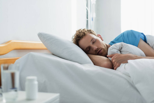 Lockenkopf-Patient schläft im Krankenhausbett neben verschwommener Flasche mit Medikamenten und Glas Wasser - Foto, Bild