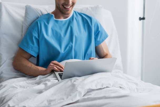 vue recadrée de pigiste heureux en robe de patient en utilisant un ordinateur portable dans le lit d'hôpital  - Photo, image