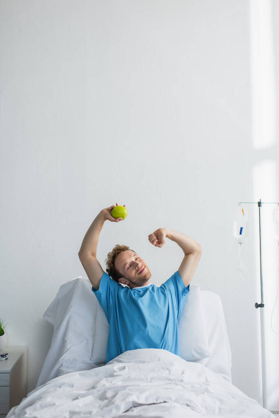 pleased man with closed eyes stretching in hospital bed and holding apple  - Fotoğraf, Görsel