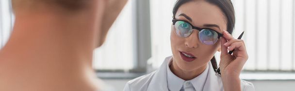brunette doctor with pen adjusting glasses while looking at blurred patient in hospital, banner - Foto, Imagen