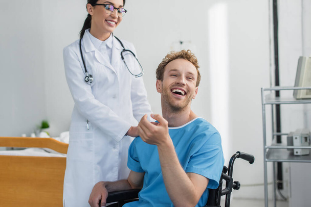 lekarz w białym płaszczu i okularach uśmiechnięty w pobliżu wesołego pacjenta niepełnosprawnego na wózku inwalidzkim  - Zdjęcie, obraz