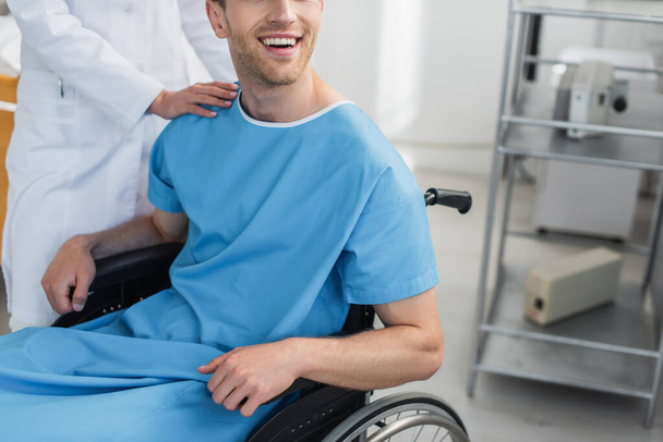 Ausgeschnittene Ansicht eines Arztes im weißen Kittel, der neben einem fröhlichen behinderten Patienten im Rollstuhl steht  - Foto, Bild