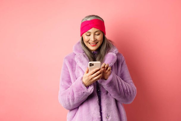 Compras en línea y concepto de moda. Moderna asiática abuelita en moda piel sintética abrigo mensaje de texto, utilizando el teléfono inteligente y mirando feliz en la pantalla, fondo rosa - Foto, Imagen