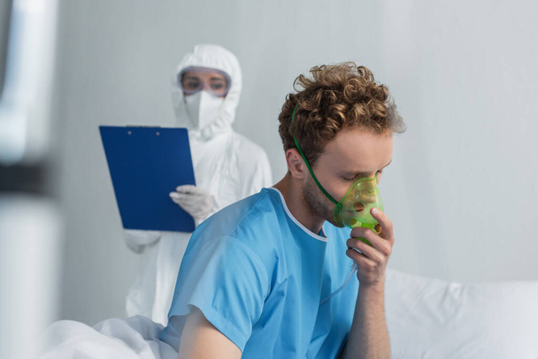 enfermo respirando la máscara de oxígeno cerca del médico borroso en el equipo de protección personal que sostiene el portapapeles  - Foto, Imagen
