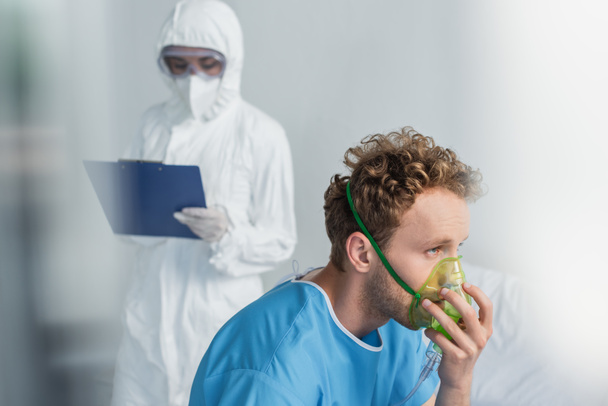 sairas potilas hengittää happinaamarissa lähellä hämärtynyt lääkäri hazmat puku pitämällä leikepöydälle  - Valokuva, kuva