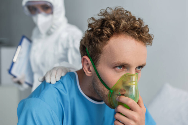 размытый доктор в защитном костюме успокаивает больного пациента, дышащего кислородной маской  - Фото, изображение