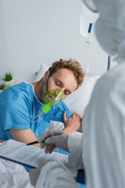 medico sfocato in tuta hazmat tenendo appunti e penna vicino al paziente in maschera di ossigeno  - Foto, immagini