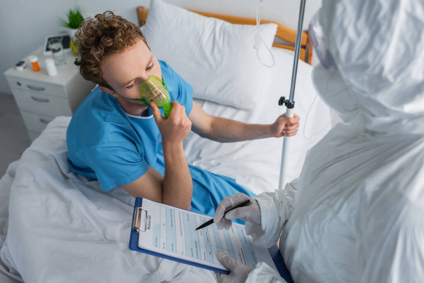 Hochwinkelaufnahme des Arztes im Schutzanzug mit Klemmbrett und Stift in der Nähe des Patienten in Sauerstoffmaske  - Foto, Bild