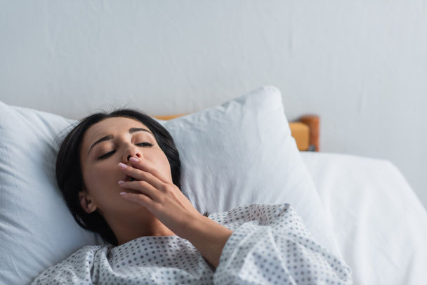 brunette vrouw in patiënt toga geeuwen terwijl liggend in ziekenhuis bed  - Foto, afbeelding