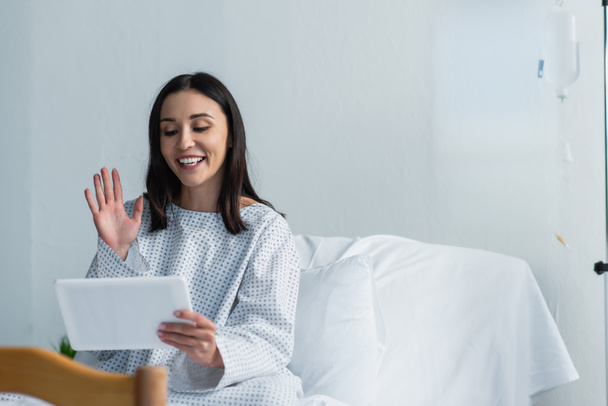 happy woman in patient gown waving hand while having video call in hospital  - Photo, Image