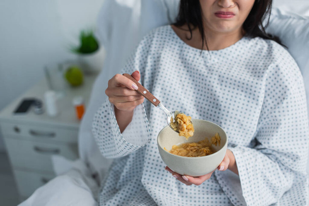 vista parcial de la mujer disgustada sosteniendo cuchara con hojuelas de maíz y tazón en el hospital  - Foto, imagen