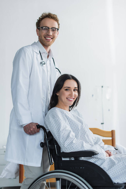 médecin joyeux en manteau blanc debout derrière femme handicapée joyeuse en fauteuil roulant  - Photo, image