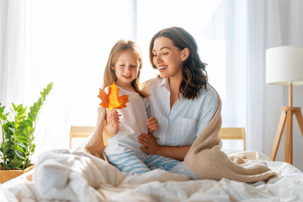 A nice girl and her mother enjoying sunny morning. Good time at home. Child wakes up from sleep. Family playing on the bed in the bedroom. - Photo, Image