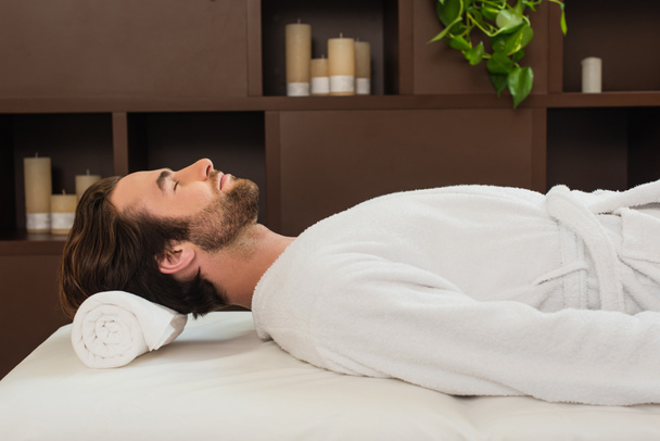 Seitenansicht des Mannes im Bademantel auf Massagetisch im Wellness-Center liegend  - Foto, Bild