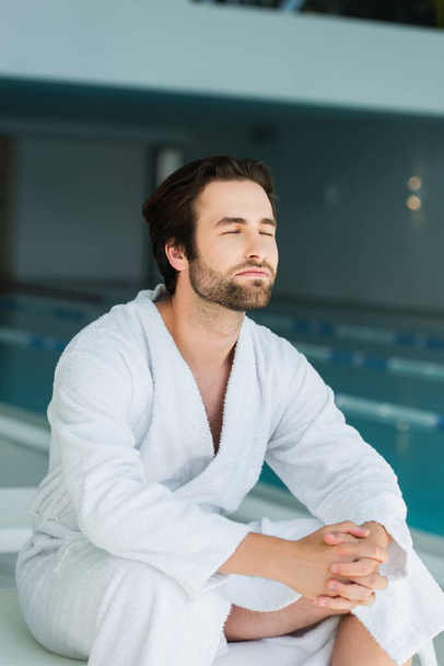 Young man in white bathrobe sitting on deck chair in spa center   - Foto, Bild