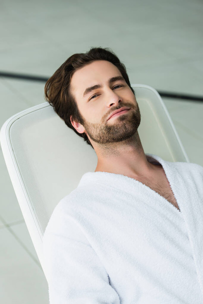 Young man in white bathrobe looking at camera on deck chair in spa center  - 写真・画像