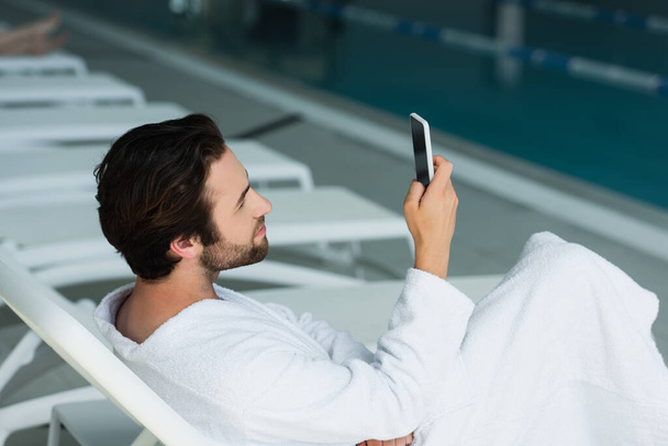Vista lateral do homem de roupão de banho usando smartphone no centro de spa  - Foto, Imagem