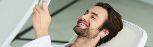 Homem alegre em fone de ouvido sem fio usando smartphone no centro de spa, banner  - Foto, Imagem